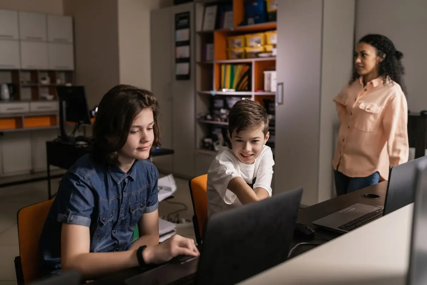 Two pupils working on laptops in a classroom while their teacher looks on